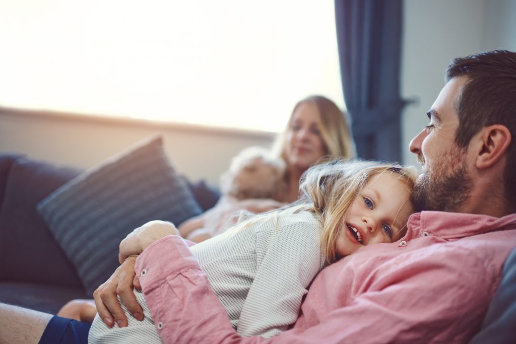 A man holds his young daughter to his chest as they relax on a sofa. In the background a woman holds another young child on the sofa.